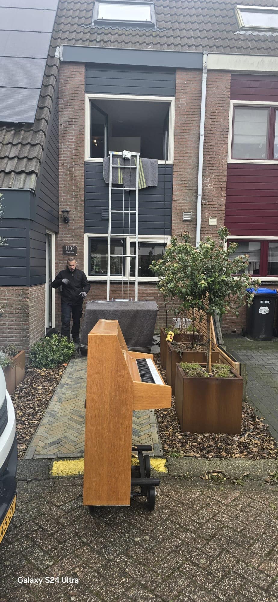 Ladderlift opgebouwd op het tuinpad naast de woning, met de bovenkant tegen het openstaande raam op de eerste verdieping en een piano op de pianosteek die omhoog gaat