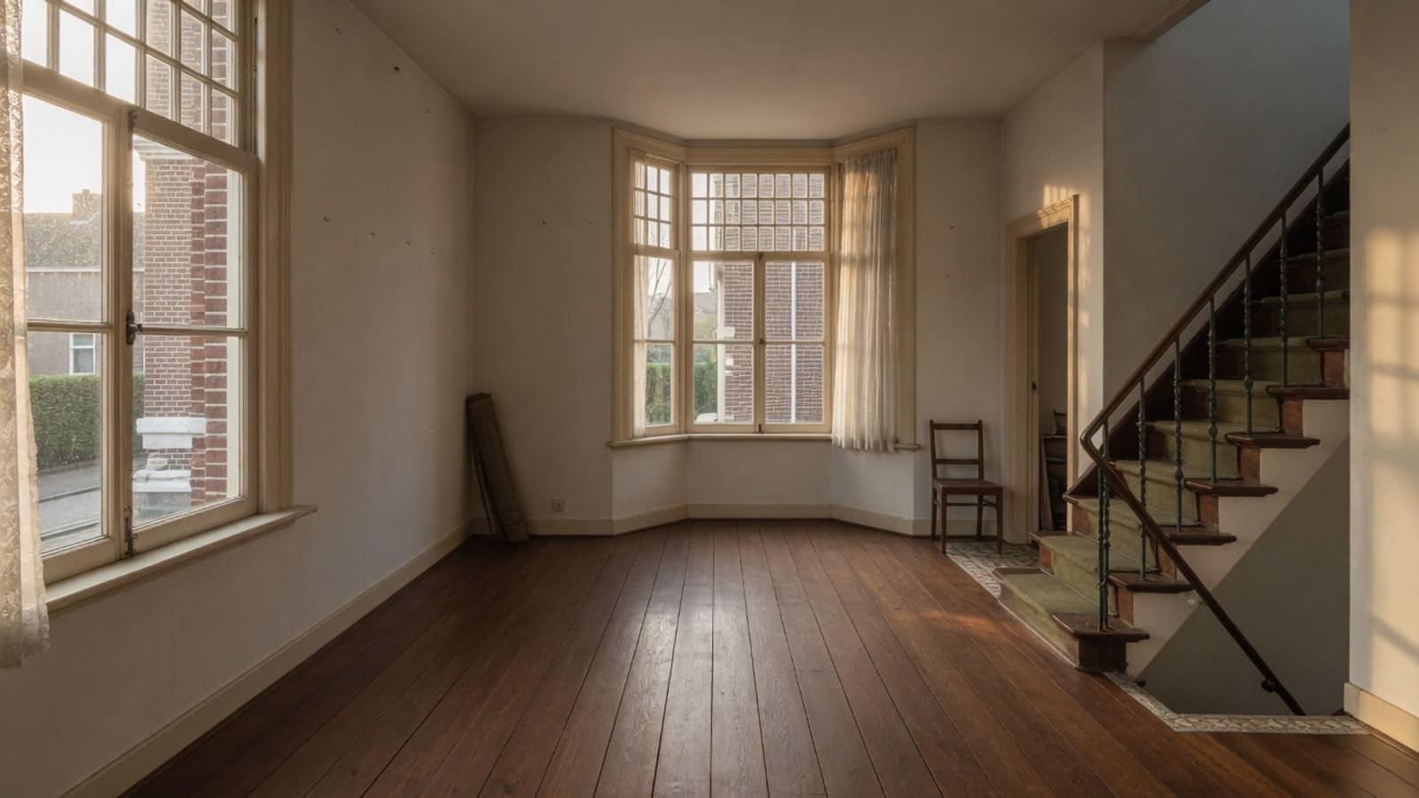 Vrijwel lege voorkamer met erker en trap, met een enkele stoel tegen de wand